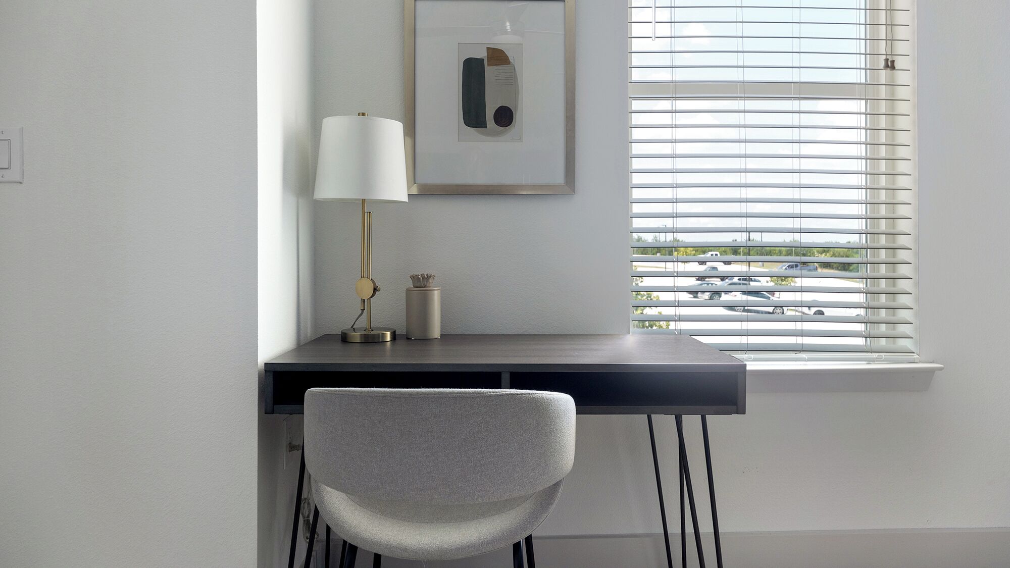 A modern desk with a lamp, a framed picture, and a chair in a room with a window and blinds.