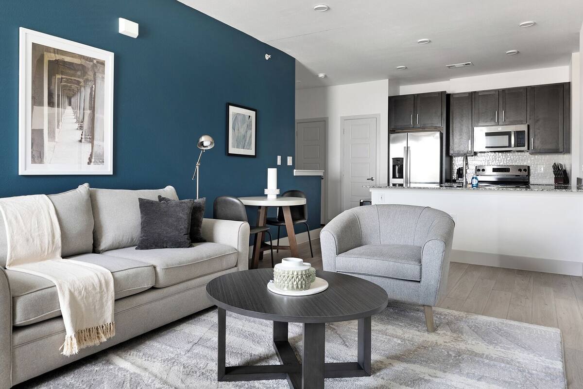 A modern living room with a grey sofa, a round coffee table, and a grey armchair. There is a kitchen area with dark cabinets and stainless steel appliances.