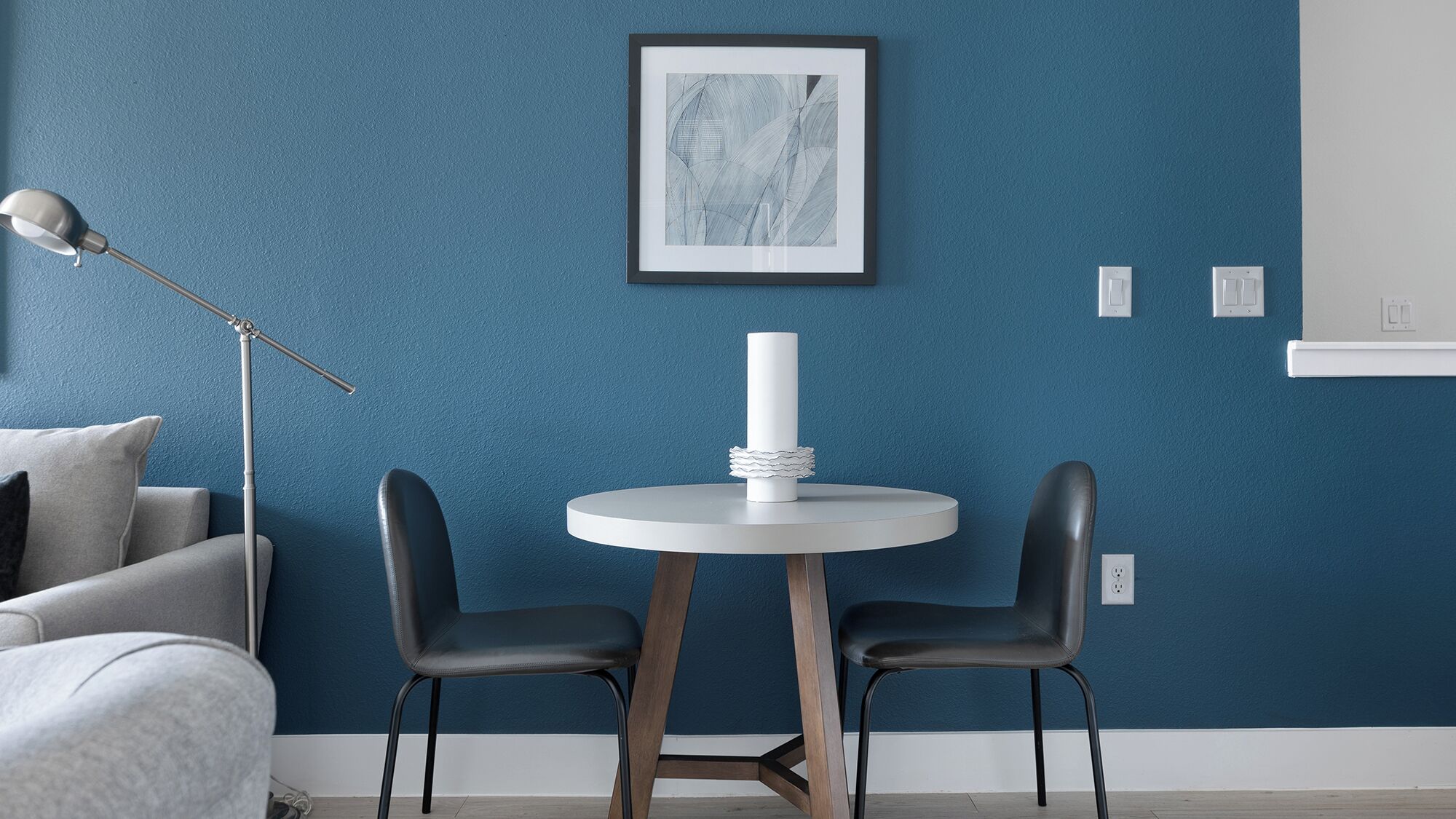 Ein Zimmer mit blauer Wand, einem runden weißen Tisch mit zwei schwarzen Stühlen, einer Stehlampe und einem gerahmten abstrakten Kunstwerk.