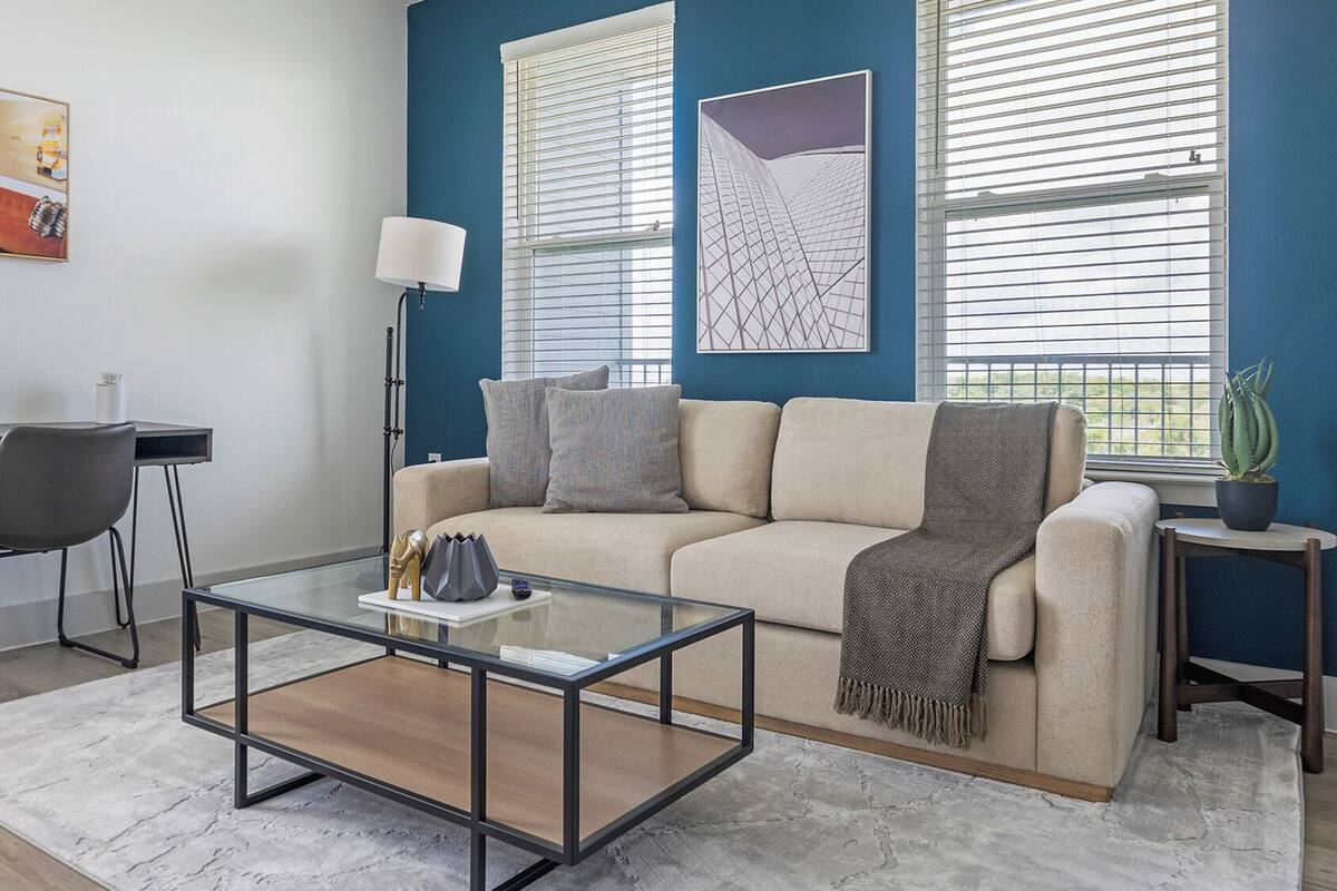 A living room with a glass coffee table, a sofa, a desk with a chair, and two windows with blinds.