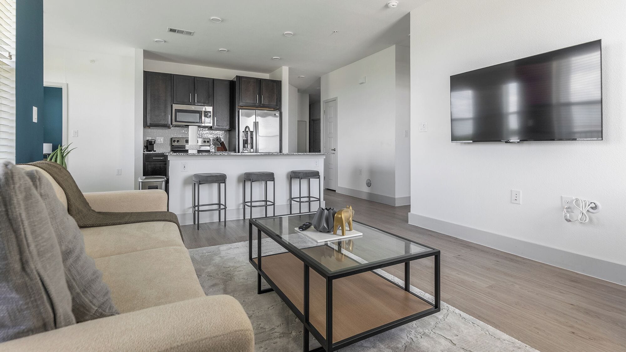 A modern living room with a glass coffee table, a beige sofa, and a kitchen in the background.