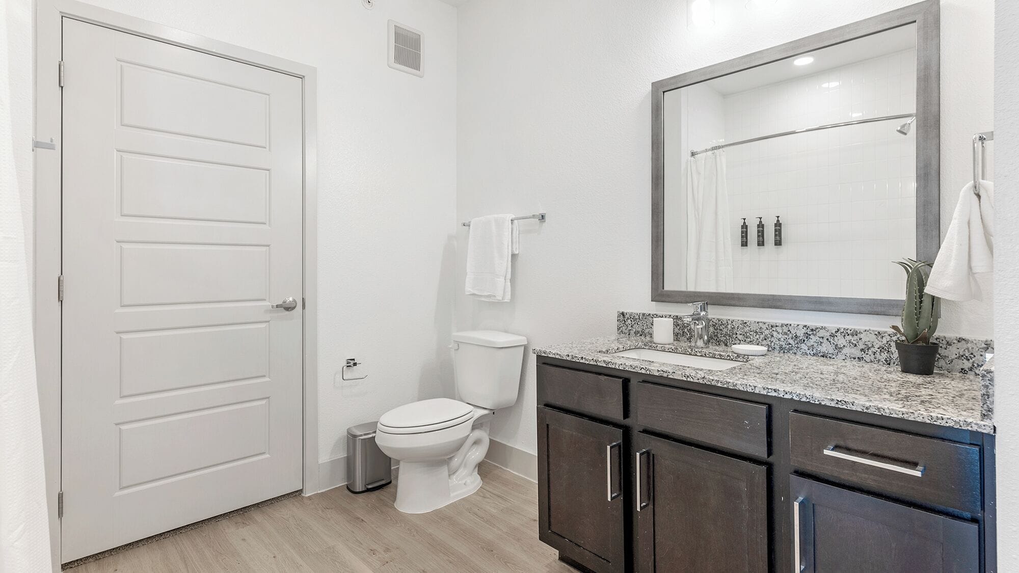 A bathroom with a white door, a toilet, a sink with a granite countertop, dark wooden cabinets, and a mirror with a towel rack.