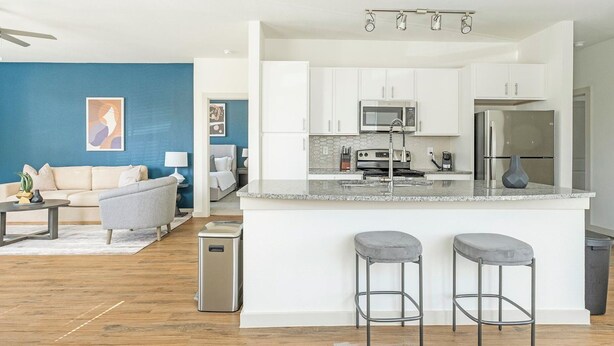 A modern kitchen with white cabinets, a granite countertop, and stainless steel appliances. There are two bar stools at the island. - Landing Apartments Waller (Waller)