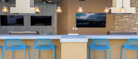 A modern outdoor bar area with a long counter, blue metal chairs, and pendant lighting.