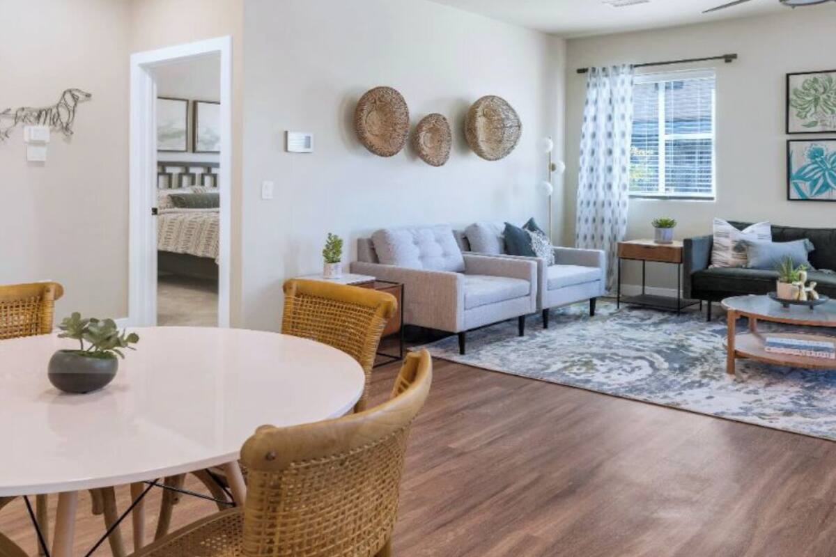 A spacious living room with a round dining table, wicker chairs, a grey sofa, and a ceiling fan.