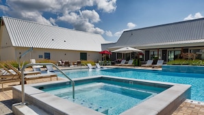 A swimming pool with lounge chairs and umbrellas, surrounded by buildings with metal roofs. - Balch Road Apartments by Landing (Madison)