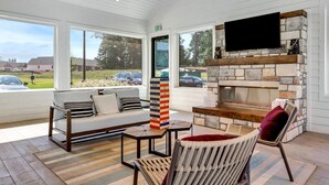 A cozy indoor seating area with a stone fireplace, a flat-screen TV, and a large window overlooking a parking lot. - Balch Road Apartments by Landing (Madison)