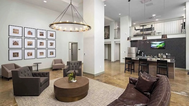 Ein geräumiges Wohnzimmer mit einer braunen Ledersofa, zwei Sesseln, einem runden Couchtisch und einer Chandelier. Im Hintergrund ist eine Küche mit schwarzem Fliesenspiegel und Edelstahlgeräten zu sehen.