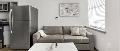 A modern kitchen with a stainless steel refrigerator, a microwave, and a stove. There is a grey sofa with cushions and a white coffee table with a plant and a small container. A framed map hangs on the wall above the sofa.