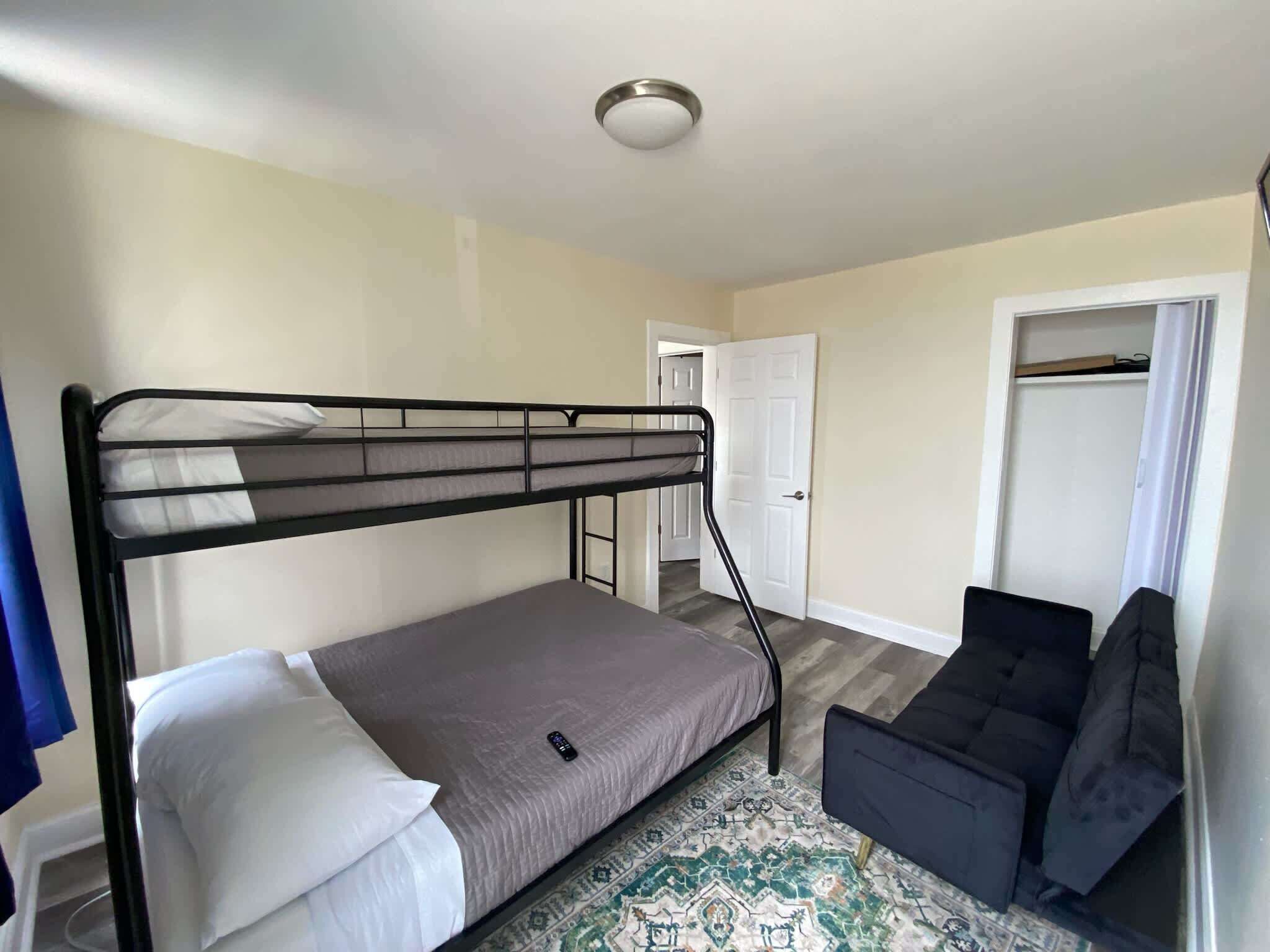 Un petit appartement d’une seule pièce comprenant un lit superposé, un petit canapé, un tapis à motifs et une armoire avec une porte.