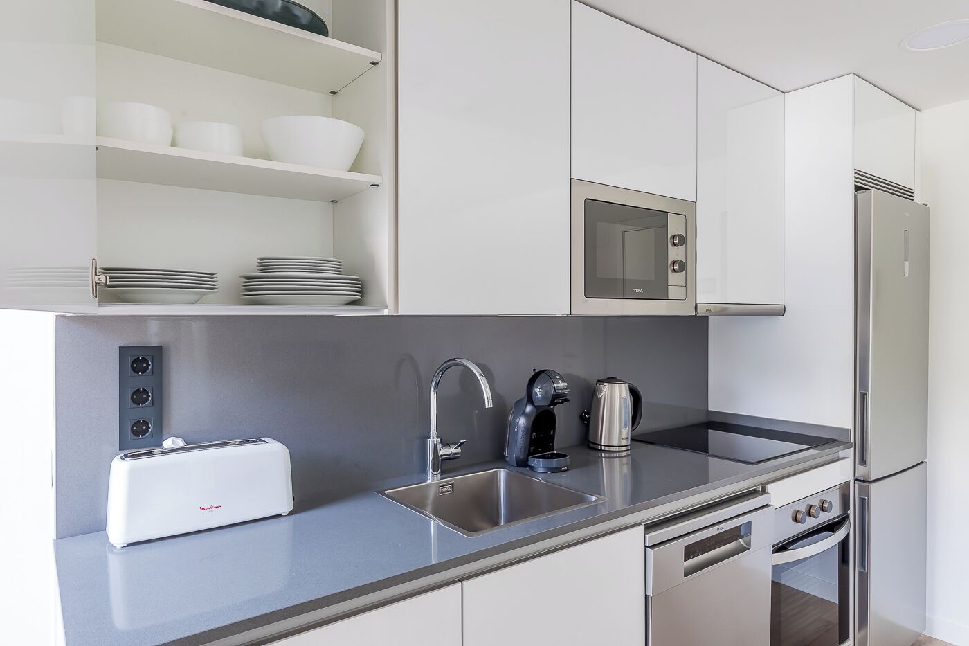 Una cocina moderna con armarios blancos, un lavamanos de acero inoxidable, un tostador, un hervidor, un microondas y un refrigerador.