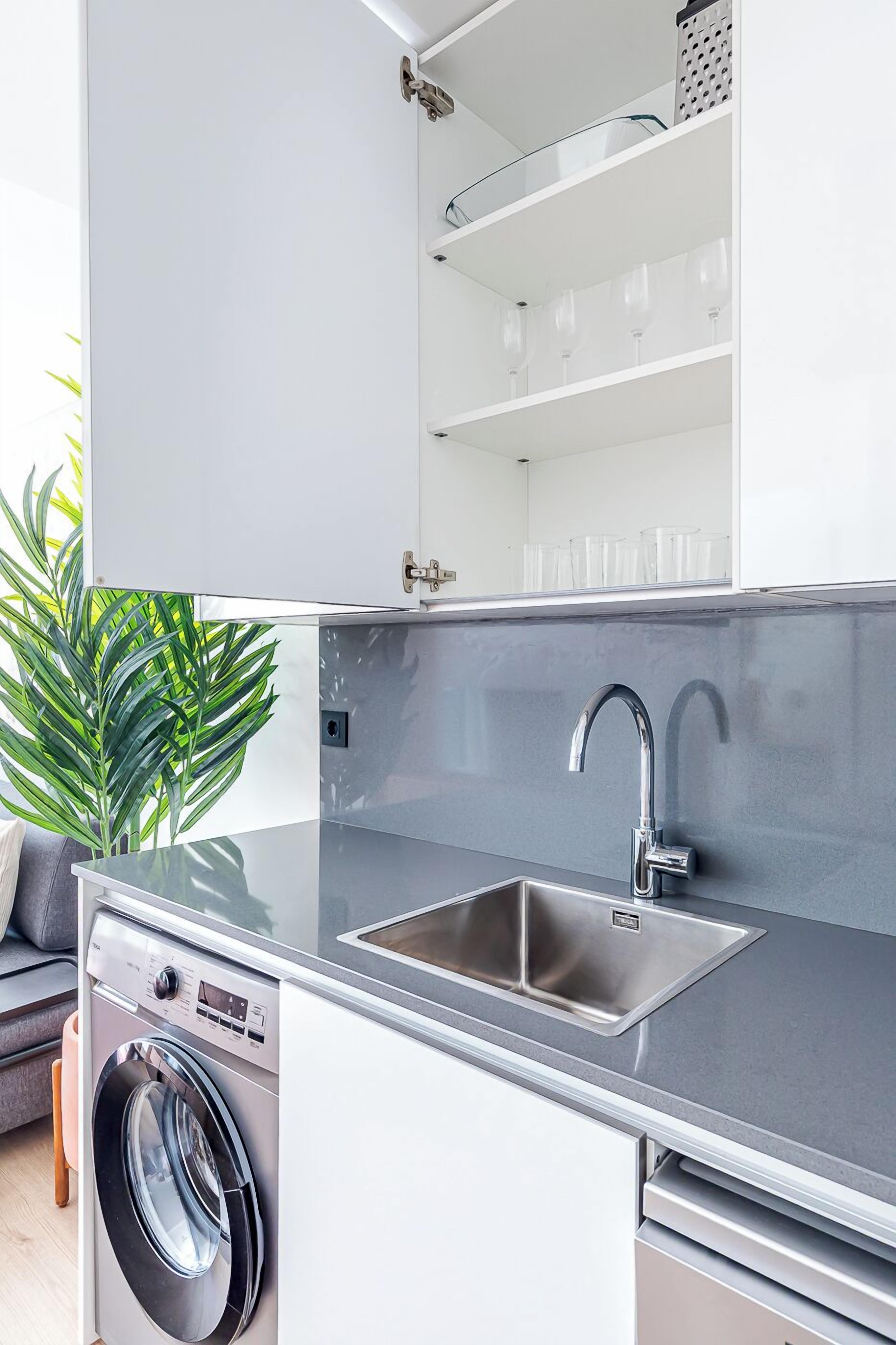 Une cuisine compacte équipée d’un lave-linge intégré, d’un évier en acier inoxydable et d’une étagère en verre au-dessus de l’évier.