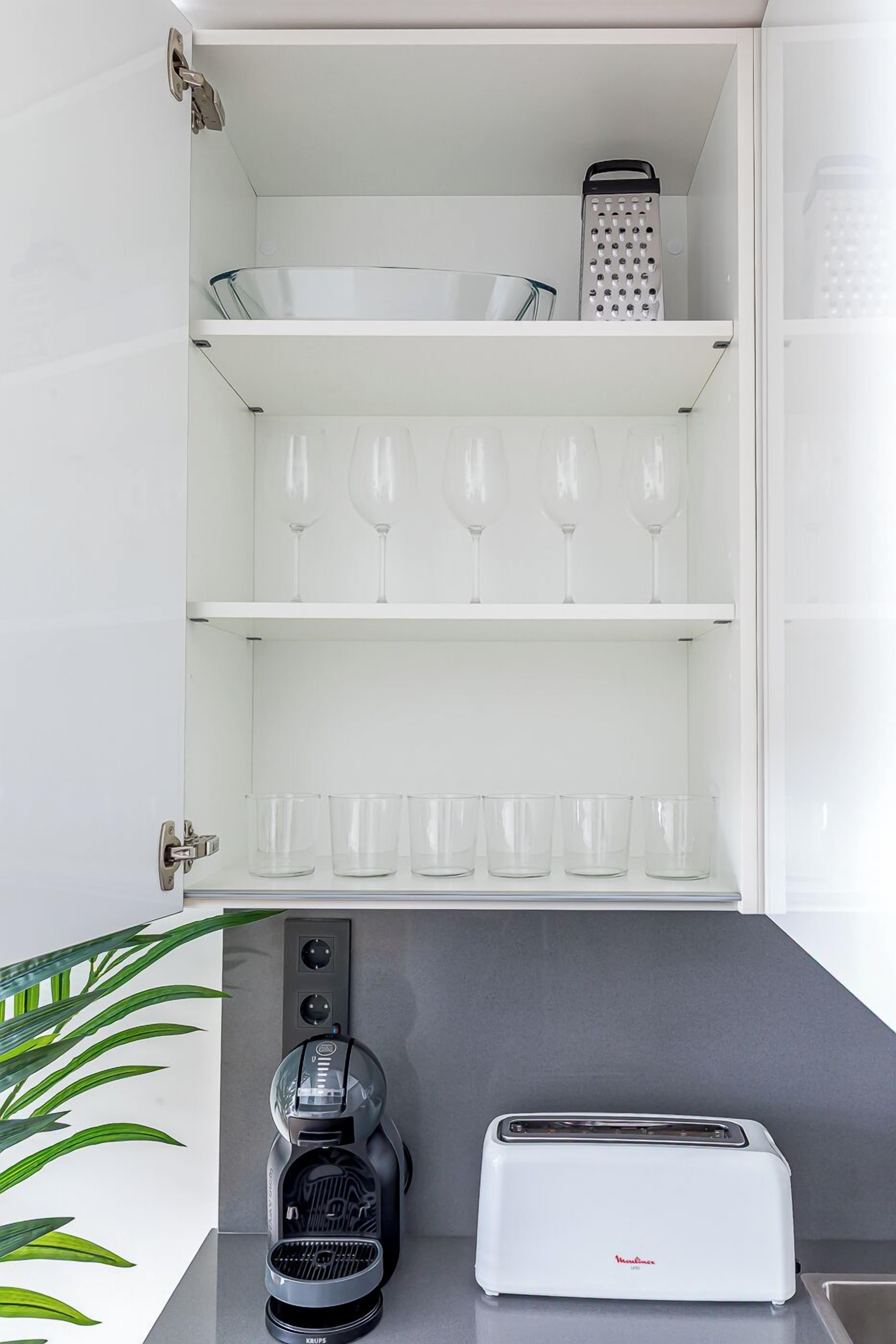 Een keuken met een witte kastinrichting met een rasp, wijnglazen en kopjes. Daaronder bevindt zich een koffiezetapparaat en een broodrooster.