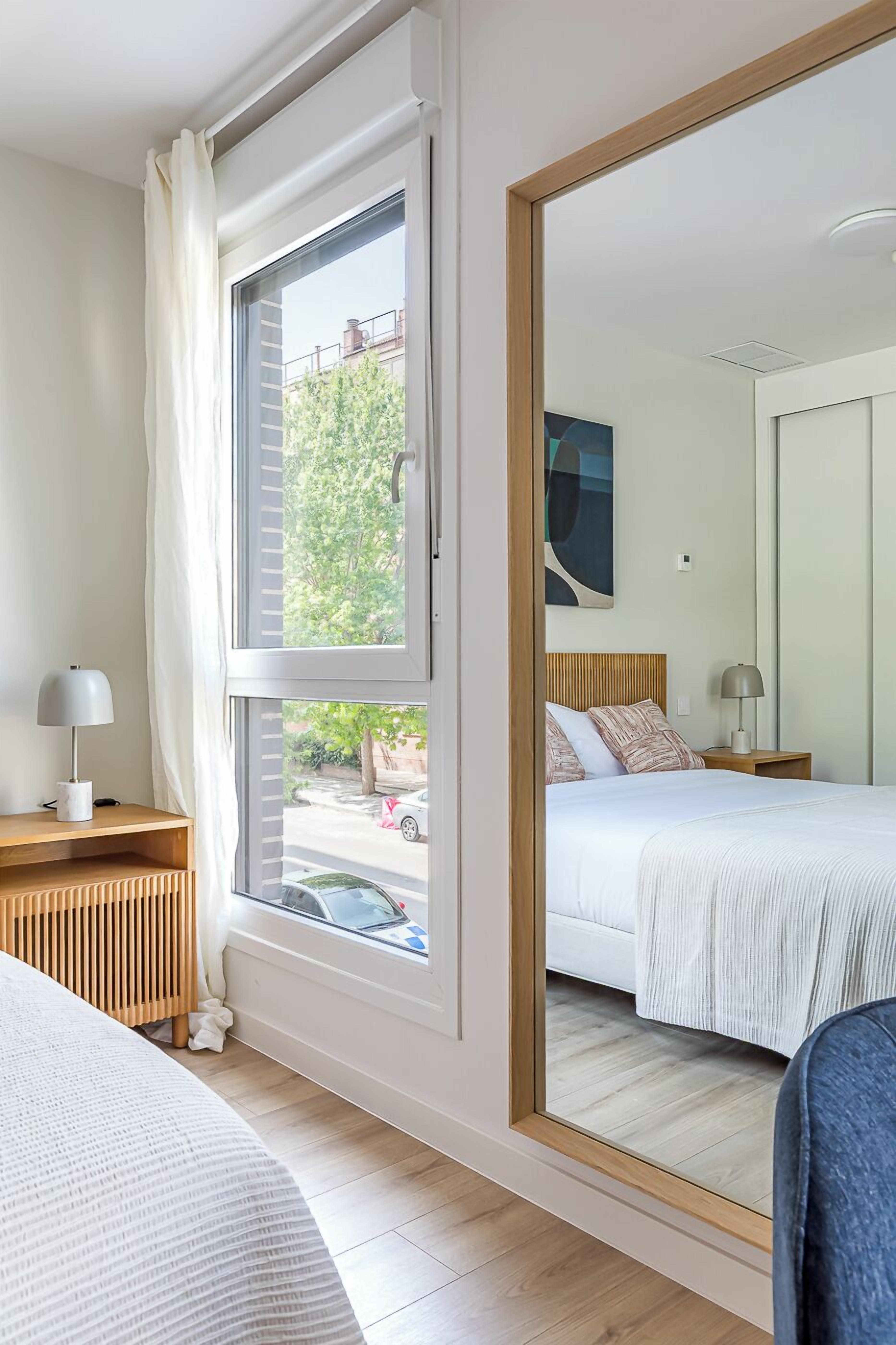 Un dormitorio con un ventanal grande, una cama con ropa de cama blanca, una mesita de noche con lámpara y un espejo que refleja la habitación.