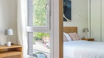 Un dormitorio con un ventanal grande, una cama con ropa de cama blanca, una mesita de noche con lámpara y un espejo que refleja la habitación.