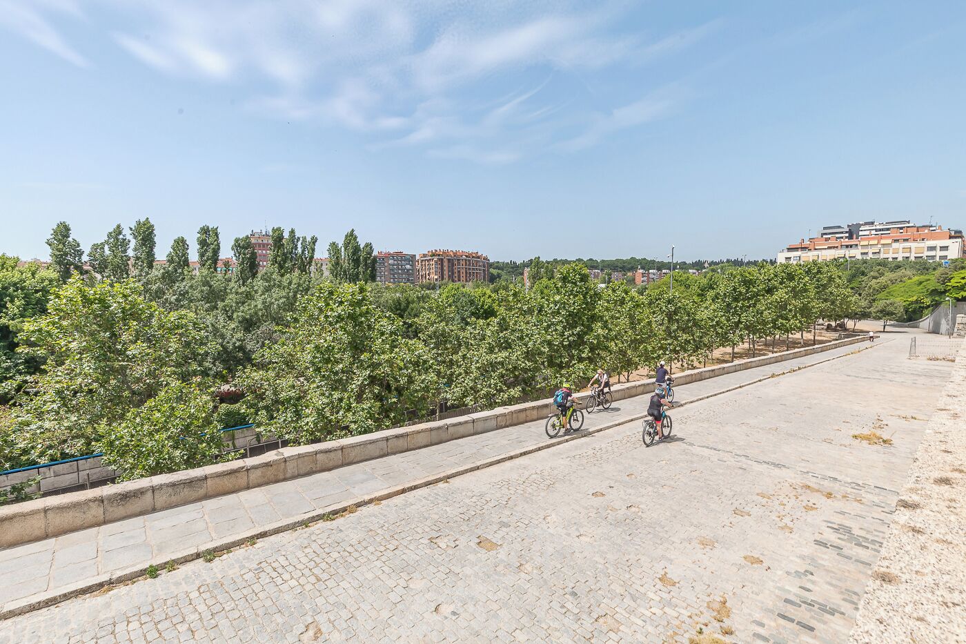 A paved pathway with cyclists, bordered by trees and buildings in the background.