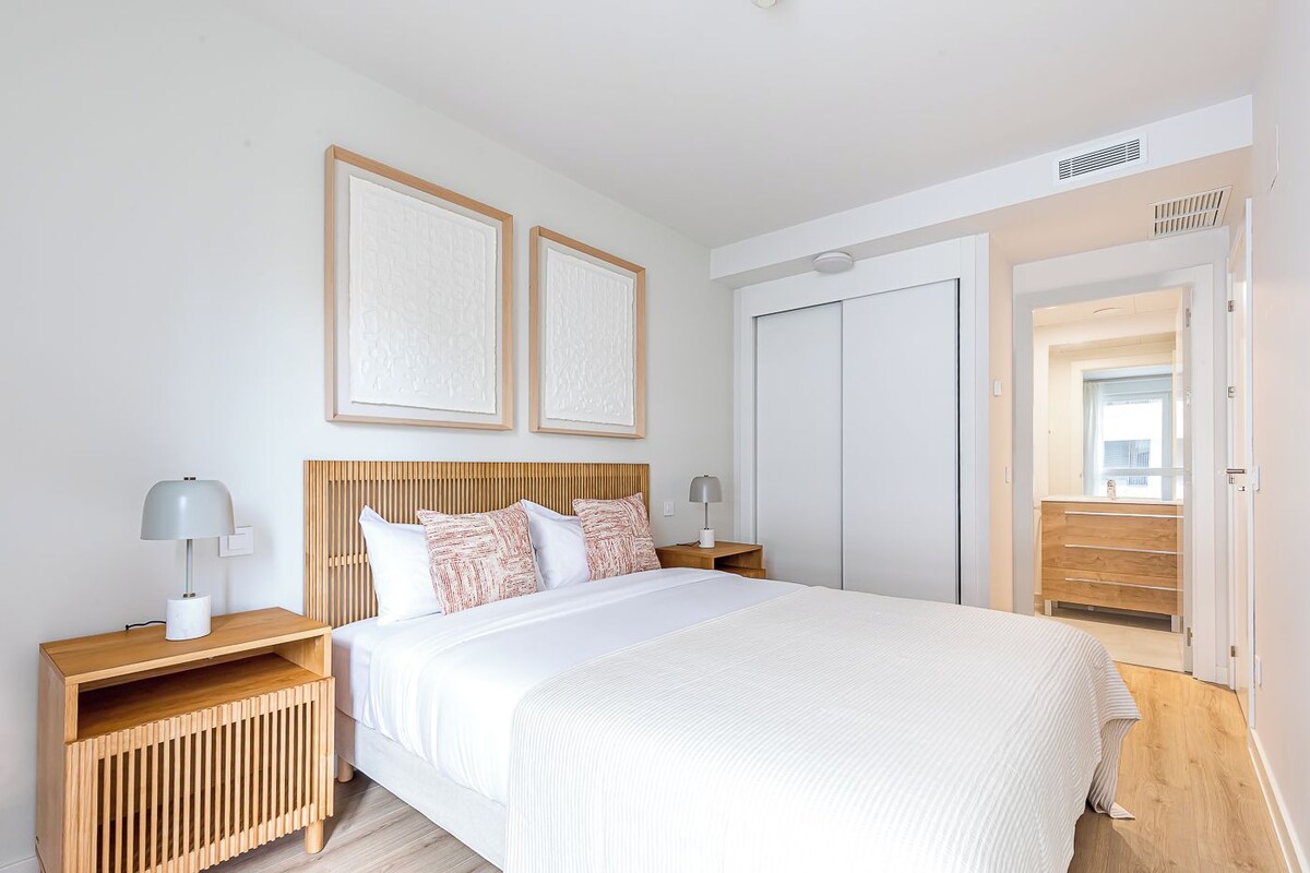 A modern bedroom with a large bed, wooden headboard, two bedside tables with lamps, a wardrobe, and a window with a view.
