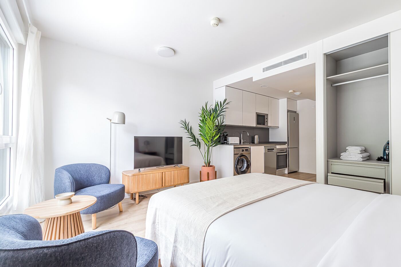 A modern hotel room with a large bed, a seating area with two chairs, a TV, a washing machine, and a kitchenette.