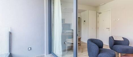 A modern apartment with a glass door leading to a balcony, a kitchen area, and two dark blue armchairs.