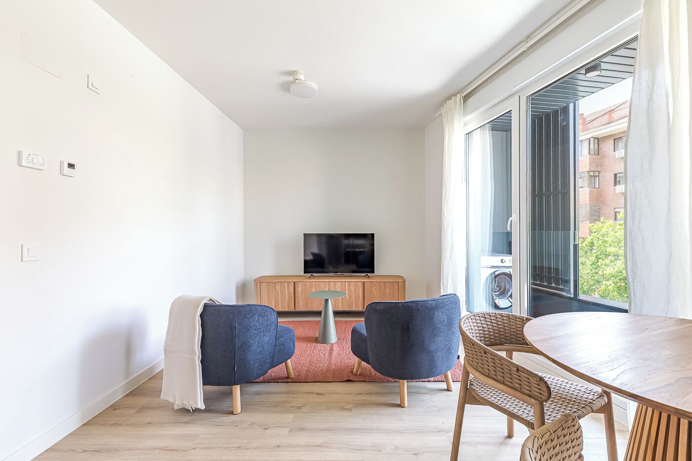 Ein modernes Wohnzimmer mit Fernseher, zwei blauen Sesseln, einem Holztisch und einem großen Fenster mit Blick auf Gebäude und Grünflächen.
