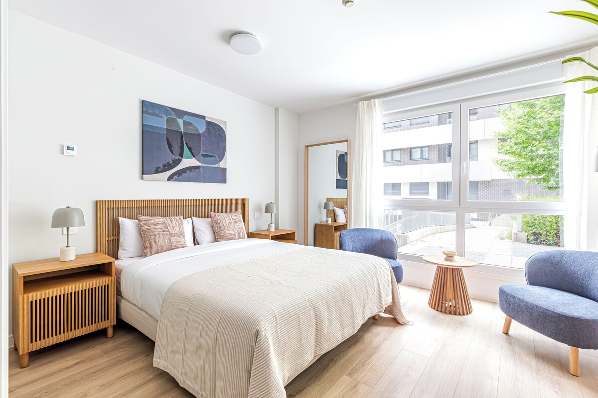 A modern hotel room with a large bed, a side table, a chair, a small table, and a window with a view of buildings and greenery.
