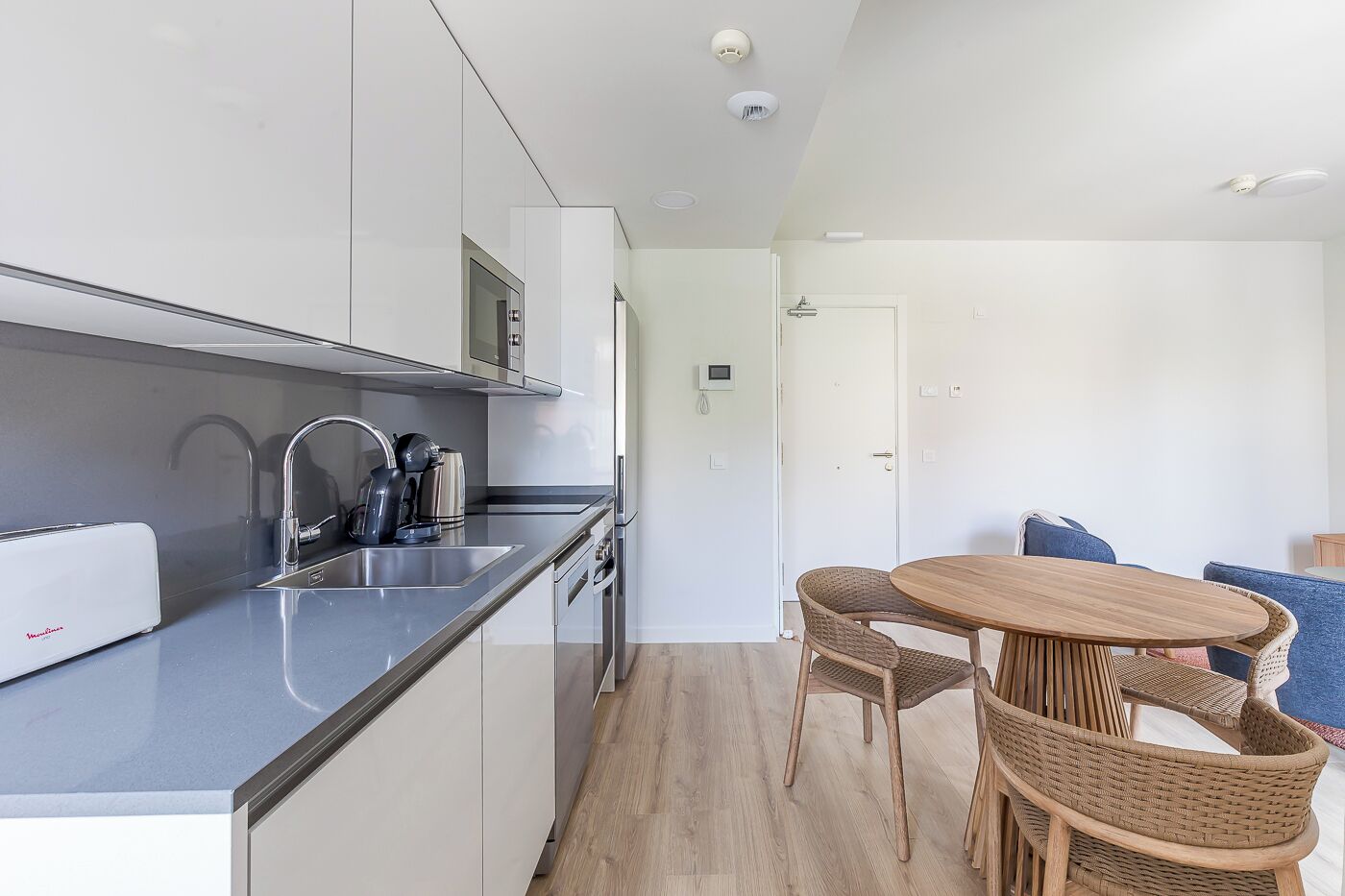 A modern kitchen with stainless steel appliances, a built-in microwave, and a coffee maker. There is a dining area with a round wooden table and wicker chairs.