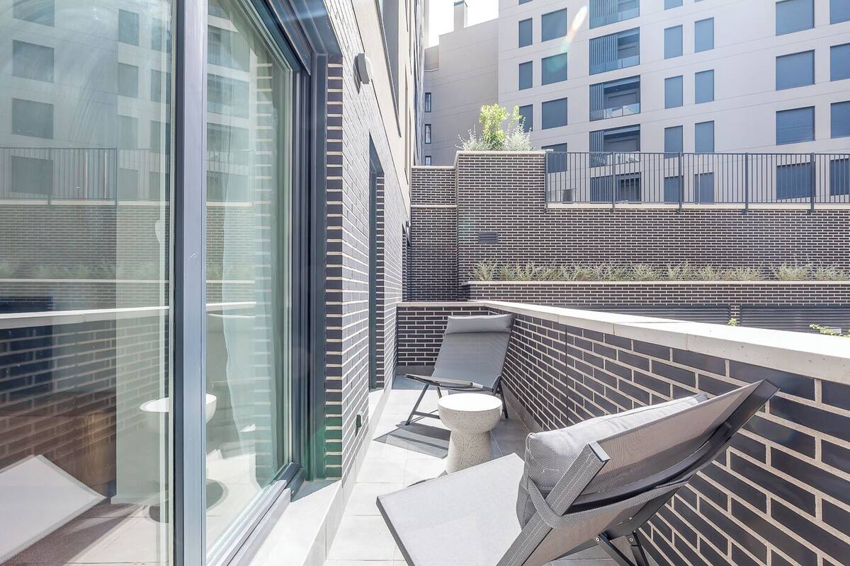A modern outdoor balcony with a glass wall, a brick wall, and a seating area with a table and chairs.
