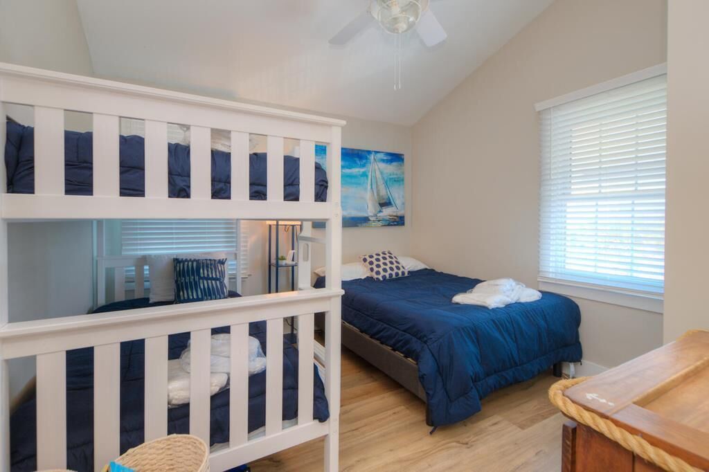 A bedroom with a bunk bed, a single bed, a ceiling fan, a window with blinds, and a painting of a sailboat.