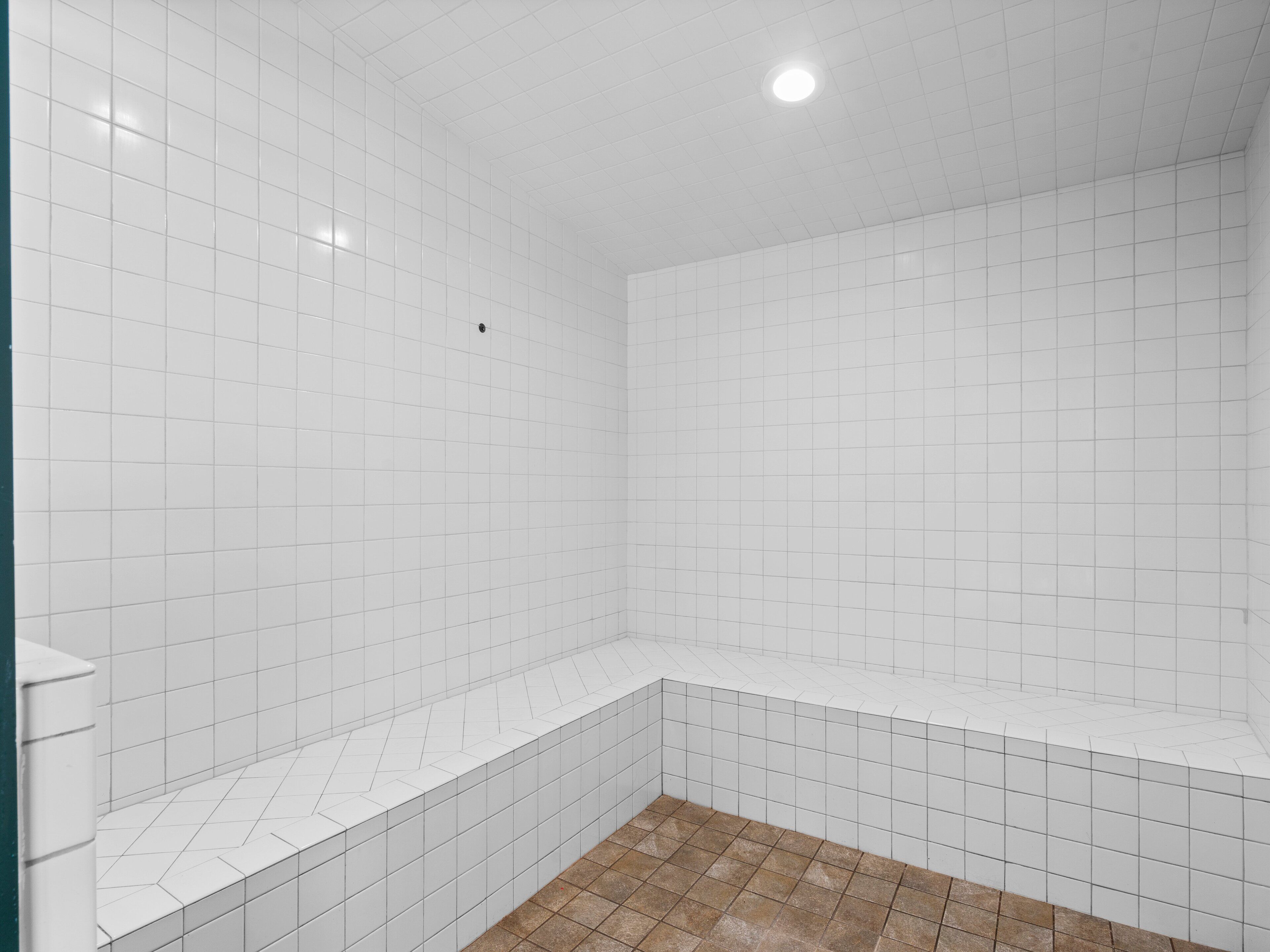 A bathroom with white tiled walls and floor, a shower area, and a corner sink.