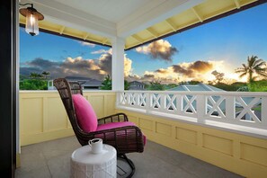 Un balcón con una silla de mimbre y una mesita, con vistas a una zona residencial con casas y palmeras.