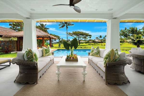 Un área de estar al aire libre techada con mobiliario de mimbre, una mesa de centro y vistas a una piscina y un paisaje tropical.