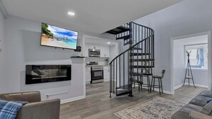 Una sala moderna con un televisor de pantalla plana, una chimenea, una escalera y una cocina al fondo.
