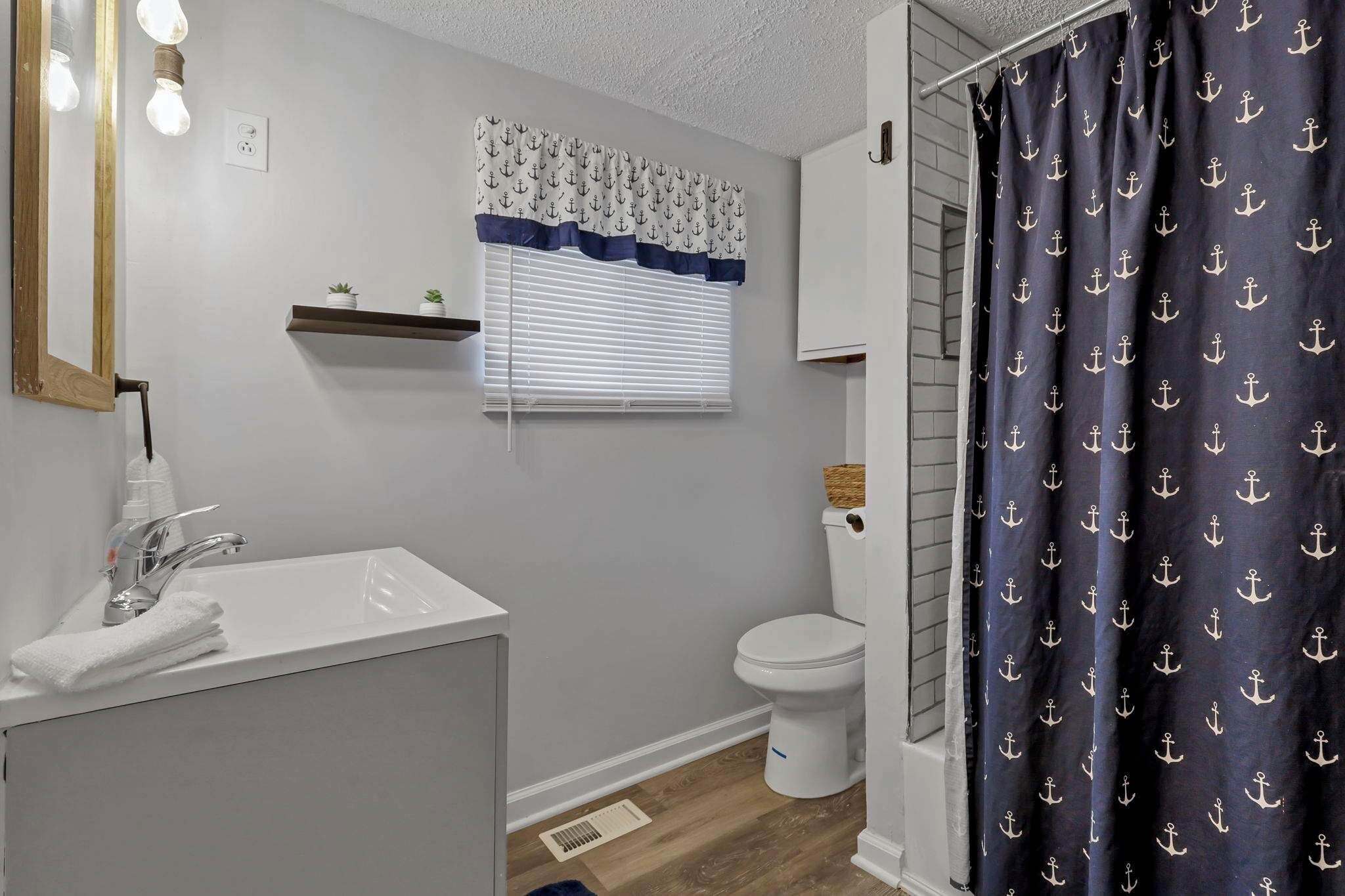 Une salle de bain avec un lavabo blanc, un miroir, une fenêtre avec un rideau à motifs et un rideau de douche orné d’imprimés d’ancres.