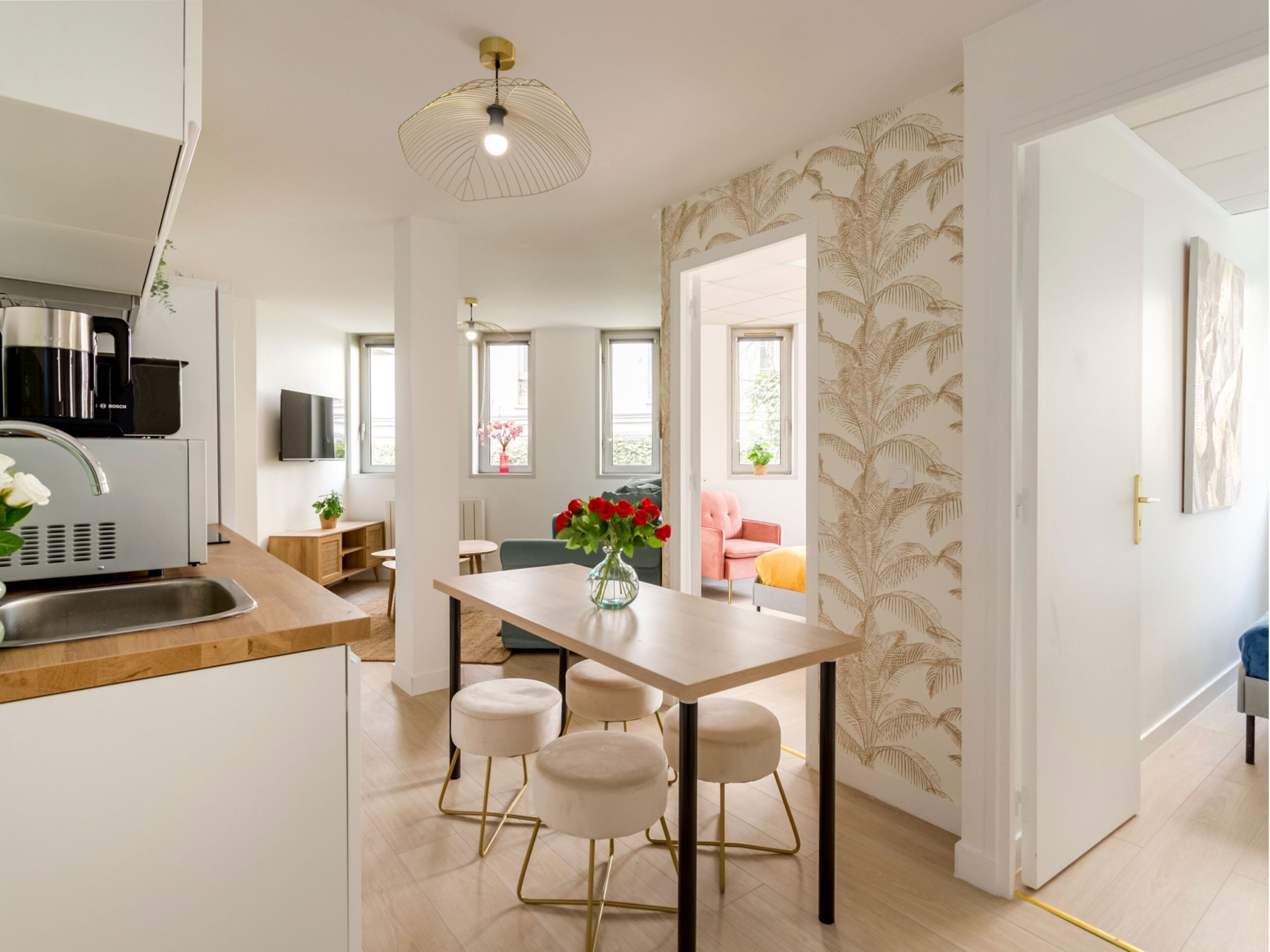 A modern kitchen with a Bosch appliance, a dining area with a vase of red roses, and a living room with a pink chair and a yellow cushion.