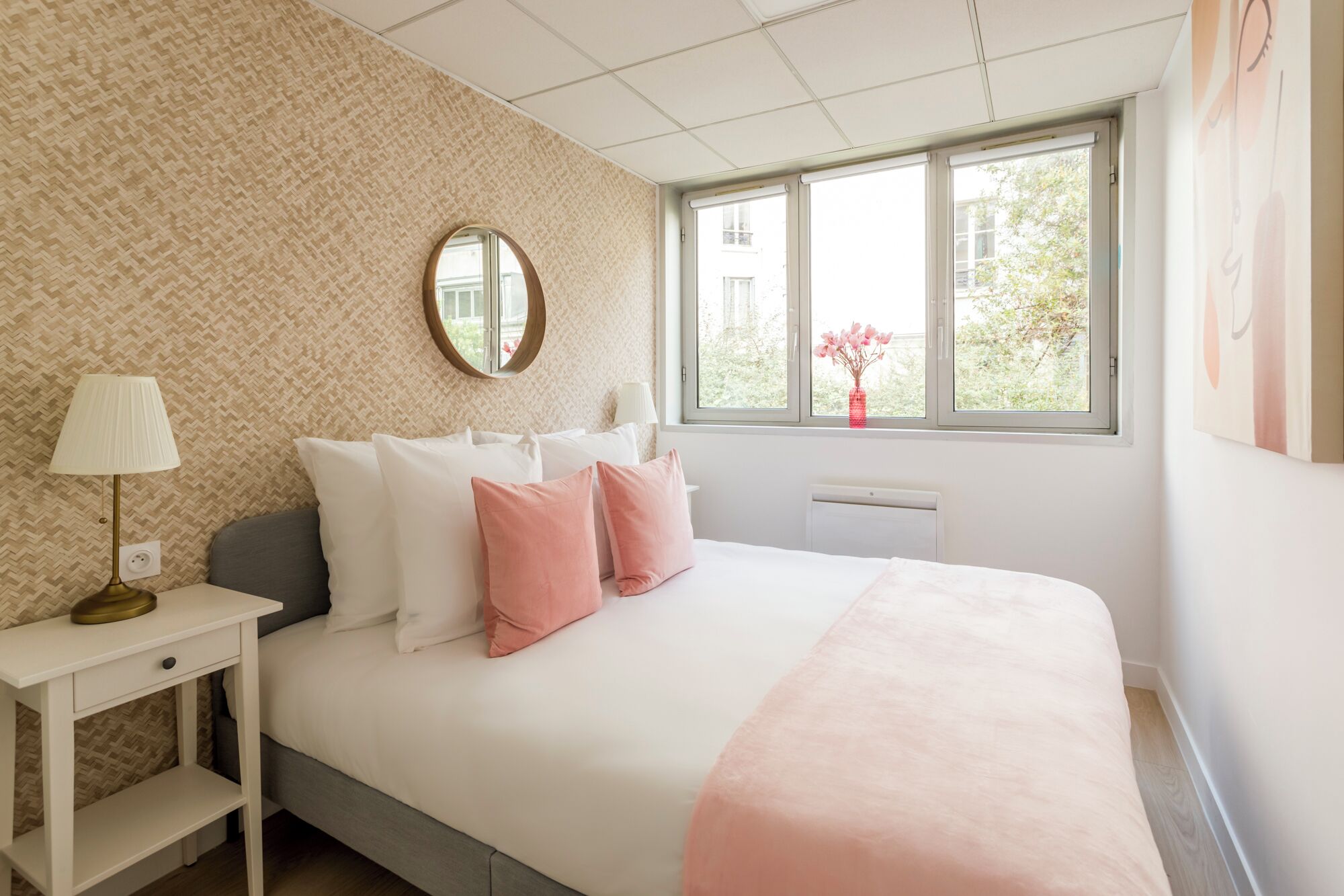 A bedroom with a bed, bedside table, lamp, and a window with a view of trees and buildings.