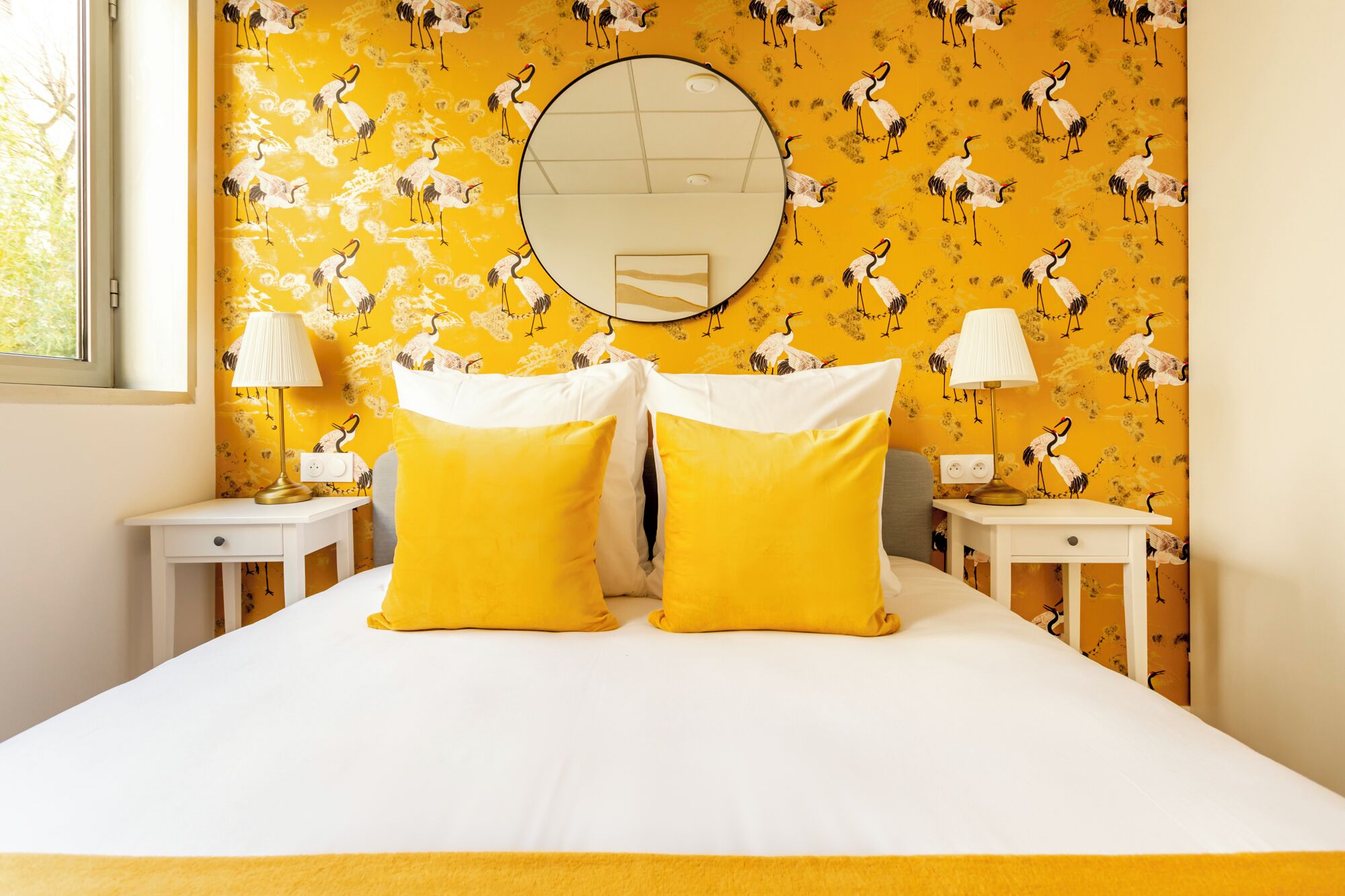 A bedroom with a bed, two bedside tables, two lamps, a round mirror, and a yellow patterned wallpaper.