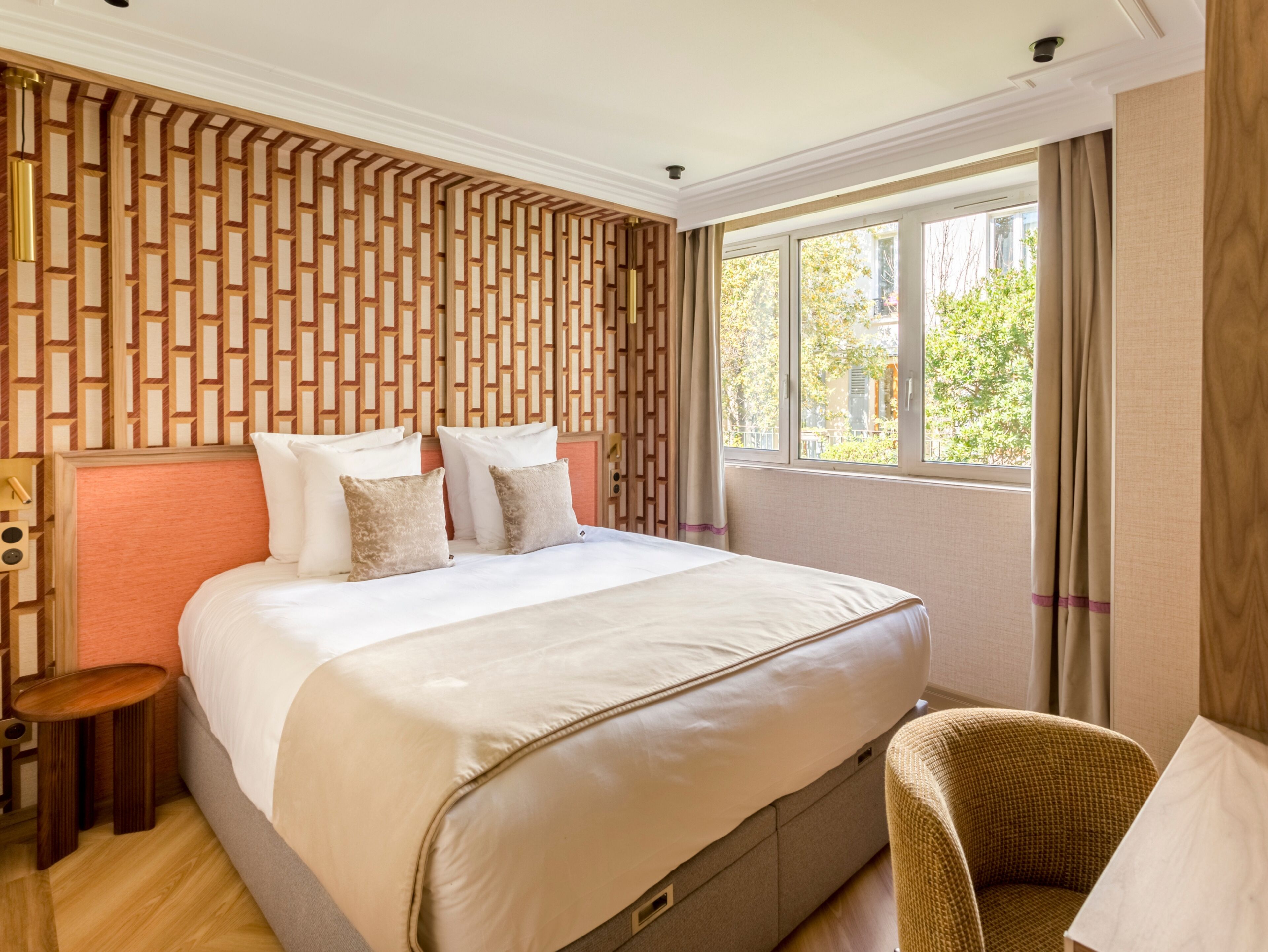 A hotel room with a large bed, a wooden headboard, a small side table, a wicker chair, and a window with curtains.