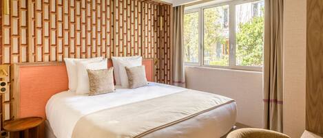 A hotel room with a large bed, a wooden headboard, a small side table, a wicker chair, and a window with curtains.