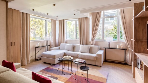 Ein modernes Wohnzimmer mit einem Ecksofa, einem Couchtisch und einer TV-Konsole. Große Fenster mit Vorhängen, ein Holzboden und ein Blick auf Grünfläche im Freien.