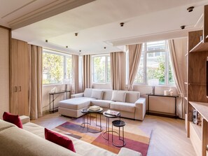A modern living room with a sectional sofa, a coffee table, and a TV stand. There are large windows with curtains, a wooden floor, and a view of greenery outside. - Family Suites - Viala I-II-IV (Paris)