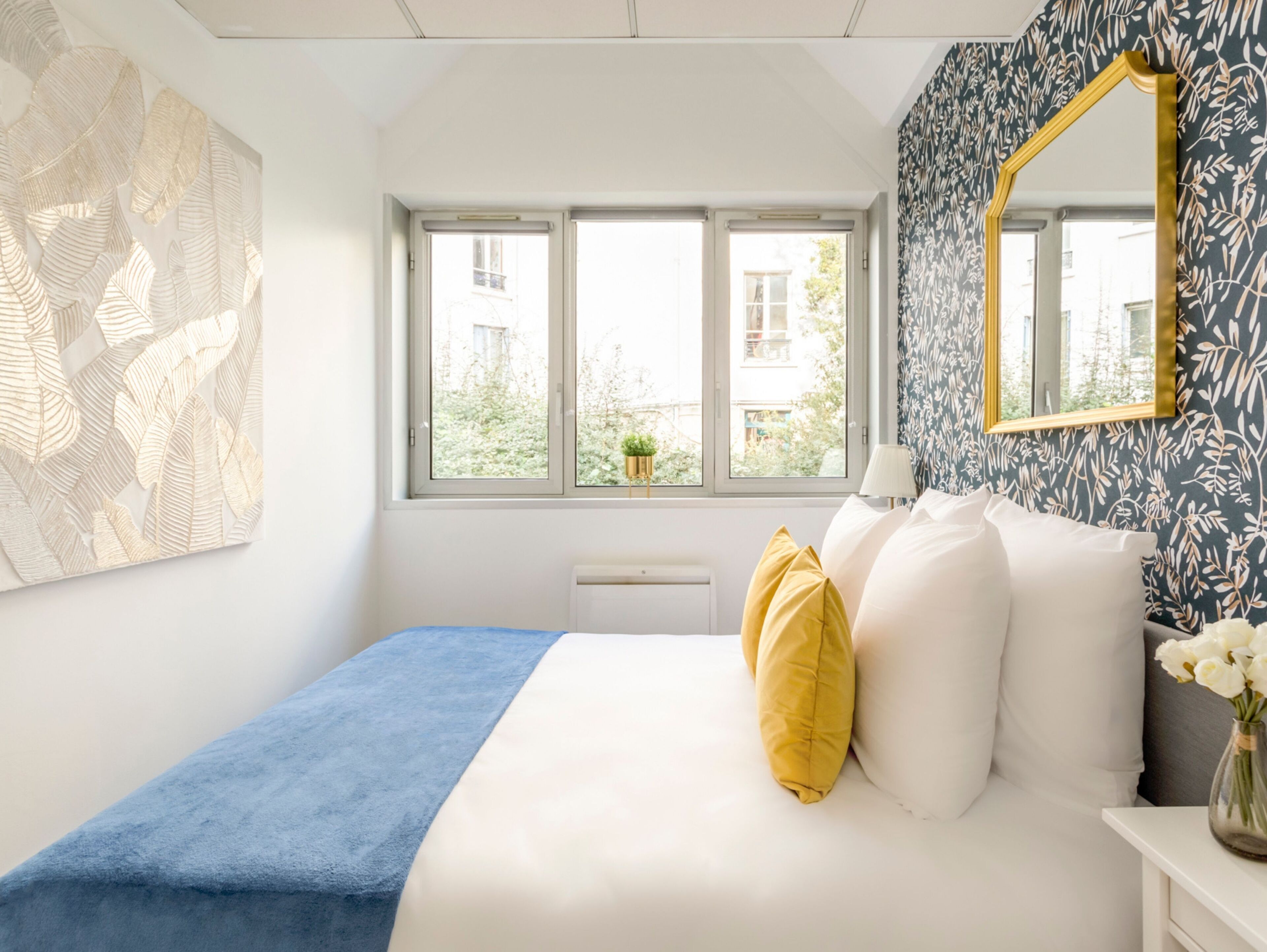 Un dormitorio con cama, ventana, espejo y pared con estampado de hojas.