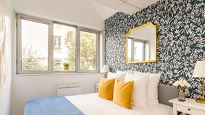 Ein Schlafzimmer mit Bett, Fenster, Heizkörper, Nachttisch, Spiegel und Wand mit Blättermotiv.