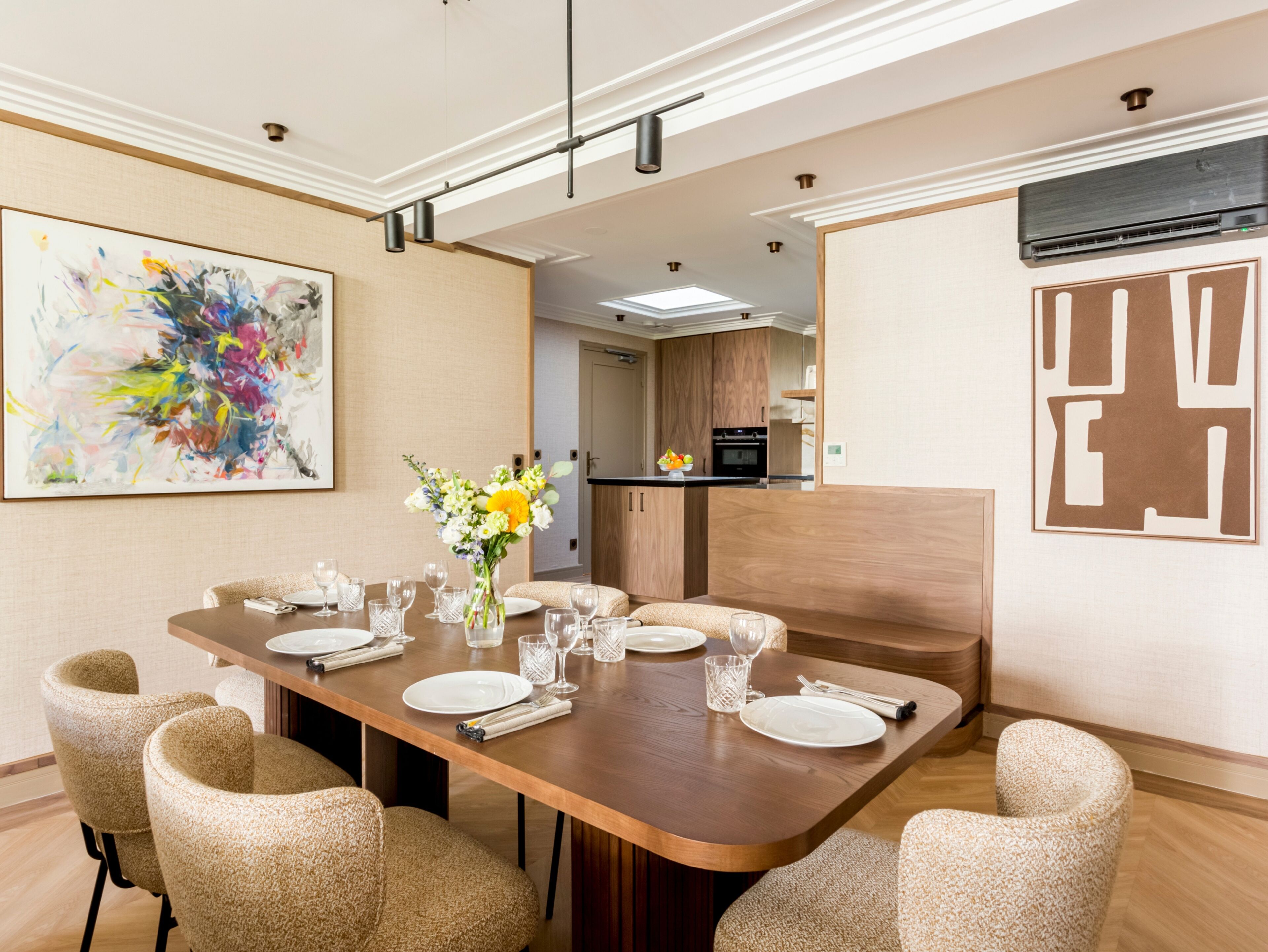 A dining area with a long wooden table set for six, surrounded by six beige chairs. There is a vase with flowers on the table, and a colorful abstract painting on the wall.