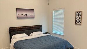 Ein Schlafzimmer mit einem Bett, einem Nachttisch, einem Fenster mit Jalousien und einem an der Wand hängenden Bild.
