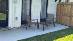 Eine überdachte Terrasse mit zwei Stühlen und einem Tisch, ein Holzzaun und ein Haus mit weißem Putz und schwarzen Fensterrahmen.