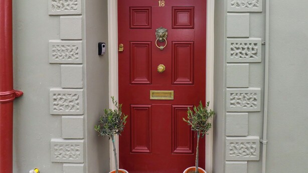 A red door with the number 18, flanked by two potted plants, and a white wall with decorative molding. - 18 Main Street (Fishguard)