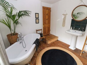 A bathroom with a freestanding bathtub, a wooden door, a small wooden stool, a white sink, and a potted plant.