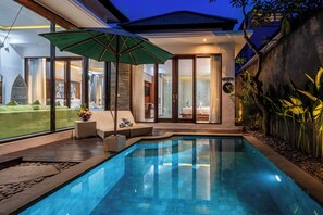Uma área de piscina moderna com uma sala de spa com paredes de vidro, um espaço de descanso com um guarda-sol verde e vista para a paisagem ao redor.