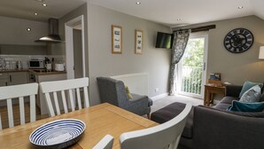 A modern kitchen with white cabinets, a microwave, and a stove. There is a dining area with a wooden table and white chairs. A gray armchair is placed near a window with a view of greenery. A gray sofa is in the living room area with a clock on the wall.