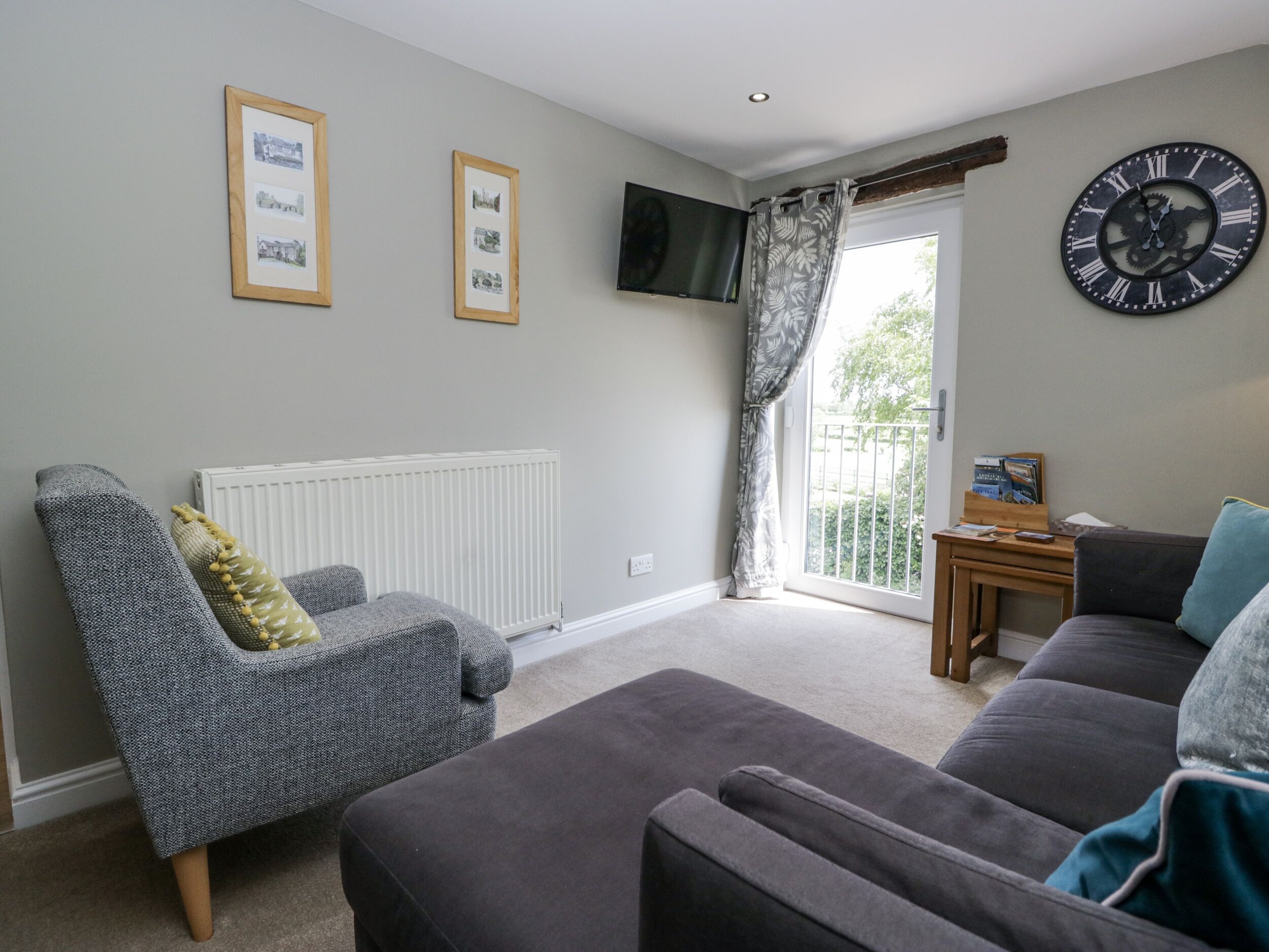 A living room with a grey sofa, a patterned armchair, a black clock, and framed pictures on the wall.